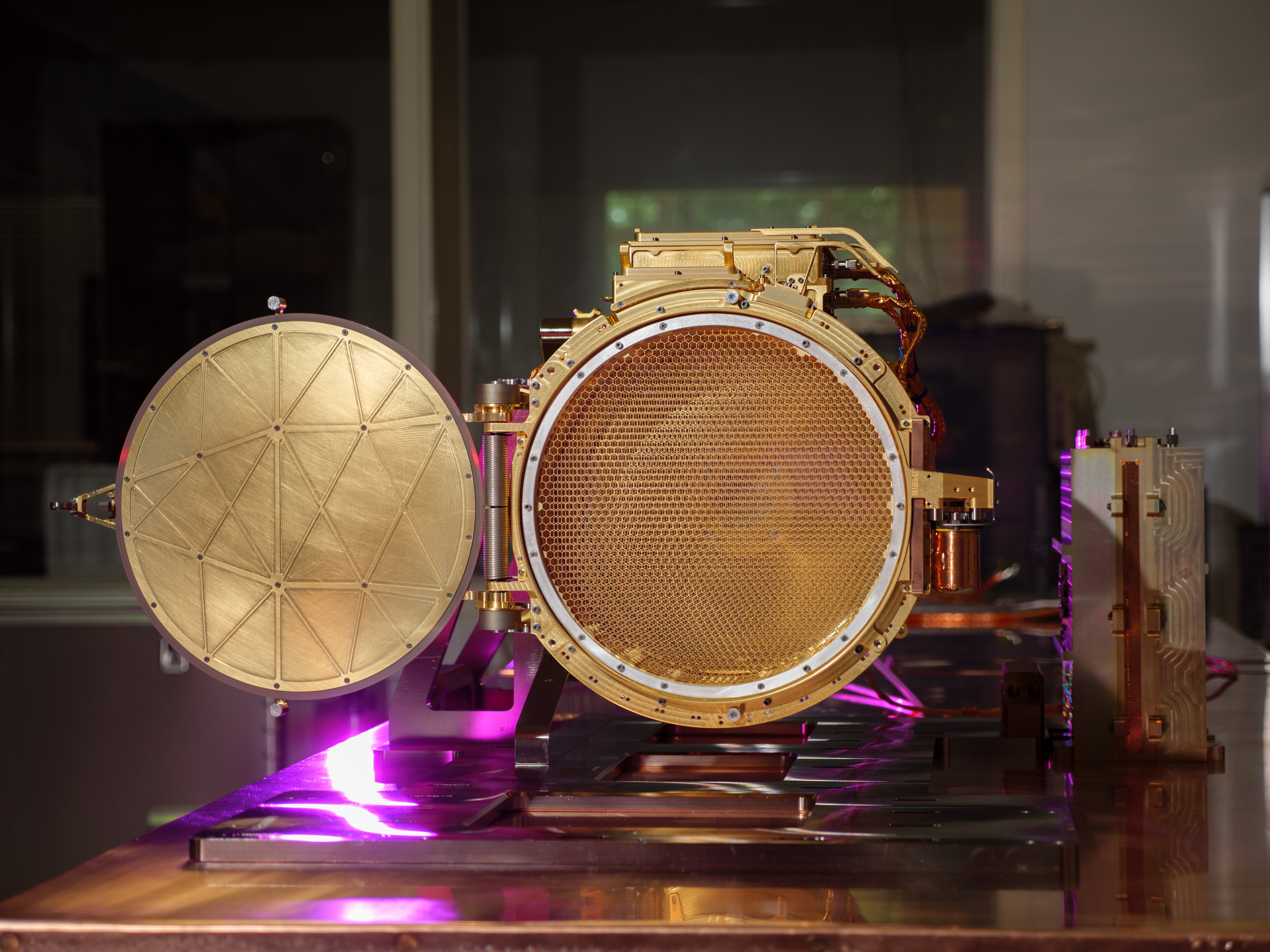 The gold colored SUDA sensor head is shown resting on a table in a clean room, seen in the center of this image. The cover to the image is shown open in the image, allowing you to see a mesh screen inside the sensor head. A purple light is seen reflecting off part of the sensor head and the table it rests on.